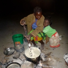 Ba Agatha talking with her daughter Gile while cooking ubwali over a traditional three-stone fire.